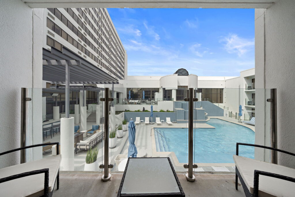 An image showing the view of the pool from one of the Cabana rooms. The balcony features a table and two white and black wicker chairs. Outside is a sunny day and the pool is a bright vibrant blue against the white of the hotel and pool chairs. There is a feature waterfall made out of bright blue tile and matching umbrellas and pillows are scattered throughout the image.
