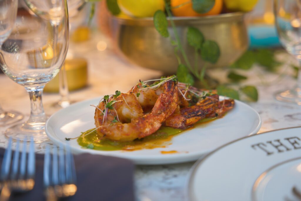 An image of fully set table with a shrimp appetizer in the center. The shrimp is bright orange, topped with micro greens, and sitting on a bed of green pesto. Around the plate is a bowl of oranges with a vine, a golden lamp, and wine glasses. The image gives off a golden warm glow and has a earthy natural feel to it.