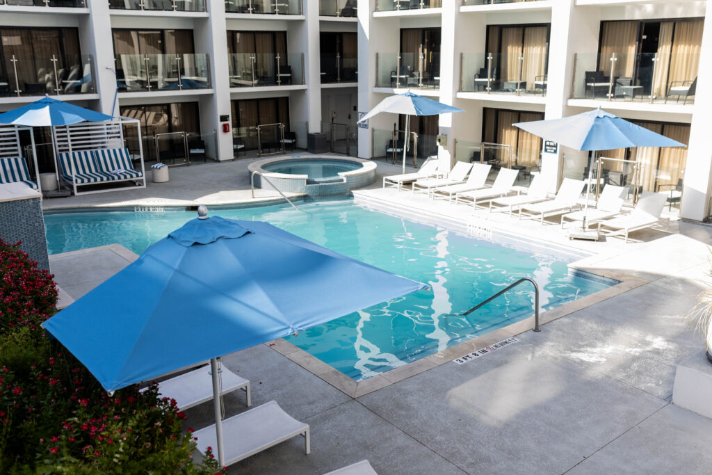 An image of the pool showcasing the balconies of the cabanas with white and black chairs. The pool is in the center of the image its square and has brigh teal water. The pool is surrounded by white lounge chairs and blue umbrellas feeling sunny and warm.