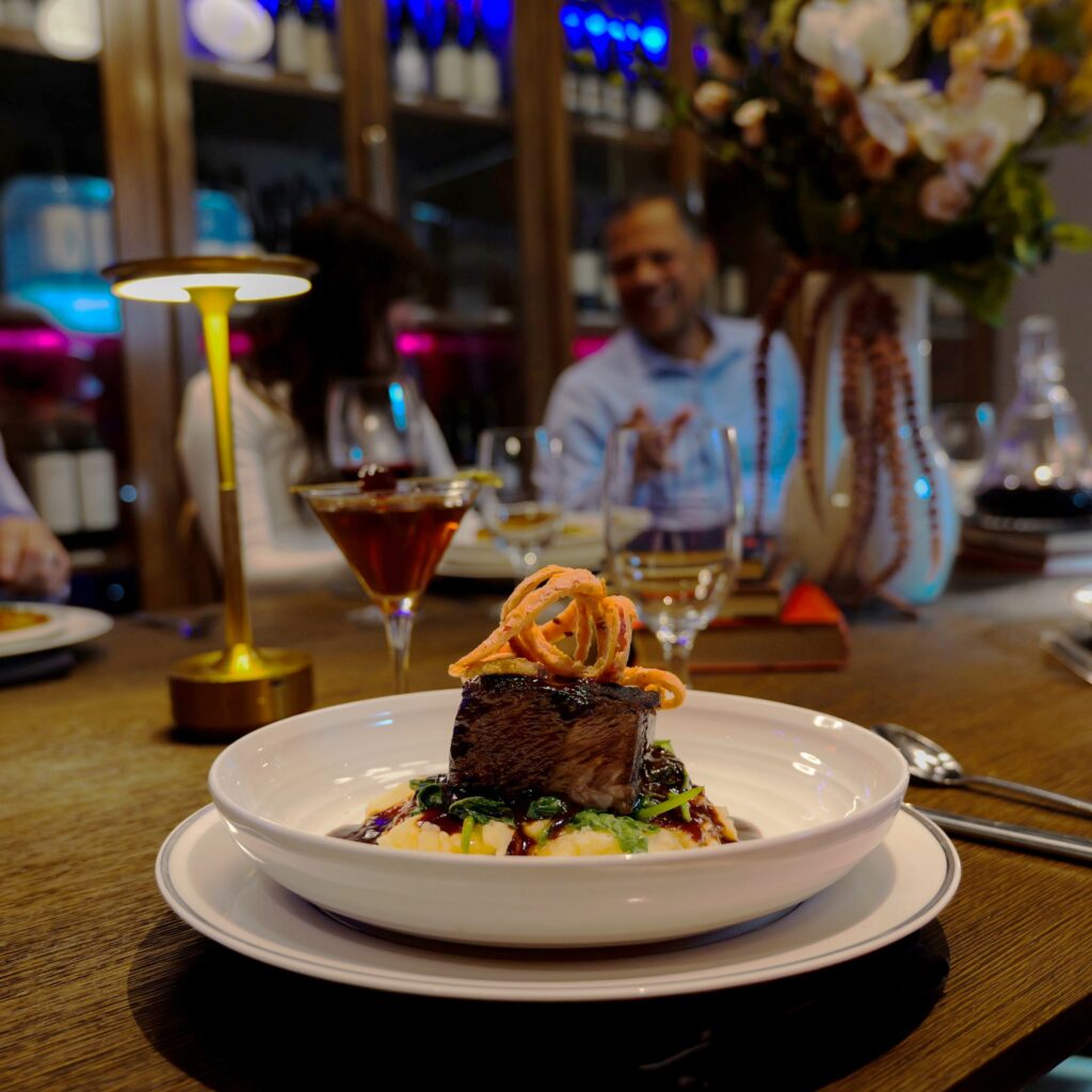 A white plate with steak and mashed potatoes is siting on a dark brown wood table. There is a cocktail next to the plate and a gold lamp. The blurred background shows a flower arrangement, colorful lights, and people sitting around the table having a conversation over food and drinks. The atmosphere is lively and pleasant with dark moody lighting.