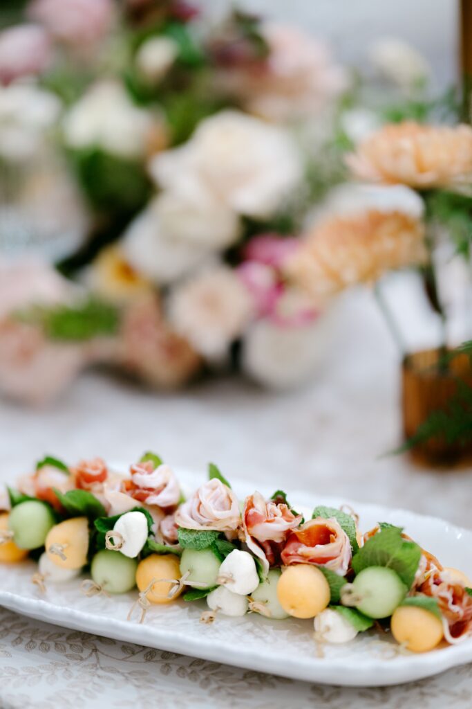 Close-up of skewered appetizers featuring melon balls, mozzarella, prosciutto, and fresh herbs, arranged neatly on a white platter. In the background, soft floral arrangements add a romantic and elegant touch.