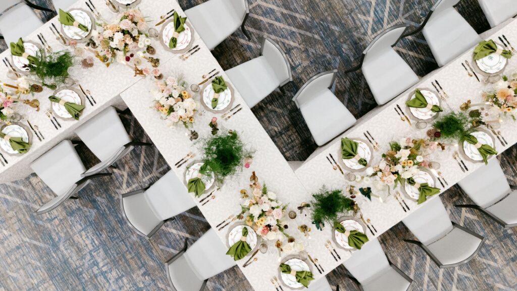 Overhead view of a U-shaped table setup featuring elegant white patterned tablecloths, fine china with green napkins, gold cutlery, and abundant floral centerpieces with greenery. The setup includes modern gray chairs arranged neatly around the table, placed on a blue and gray patterned carpet.