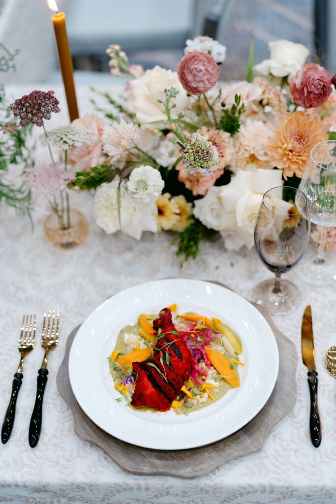 A beautifully plated gourmet dish is served on a white plate, featuring vibrant colors with roasted meat, fresh vegetables, and garnishes. The elegant table setting includes black and gold flatware, crystal glassware, and a floral centerpiece with pastel roses and dahlias. A lit candle adds a romantic ambiance, making the setting ideal for a wedding or upscale dining event.