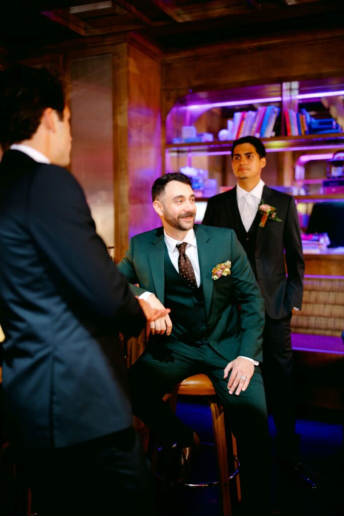 A groom in a green suit sits on a wooden barstool, smiling while interacting with two groomsmen in dark suits. The group is in a warmly lit lounge area with a wooden interior and illuminated bookshelves in the background.