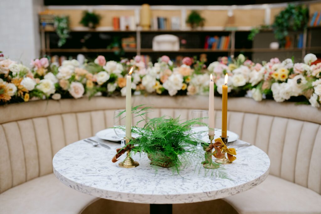 A round marble-top table set for two with elegant candles, greenery centerpiece, and fine dining place settings. The cozy booth seating is decorated with a lush floral arrangement in soft pastel colors, creating a romantic and intimate atmosphere perfect for a wedding or special occasion.
