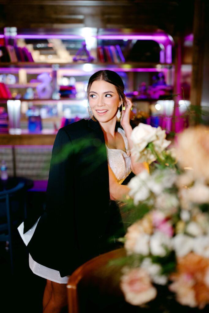 Young lady in white dress with black coat over her shoulders sitting at a bar in a colorfully lit speakeasy.