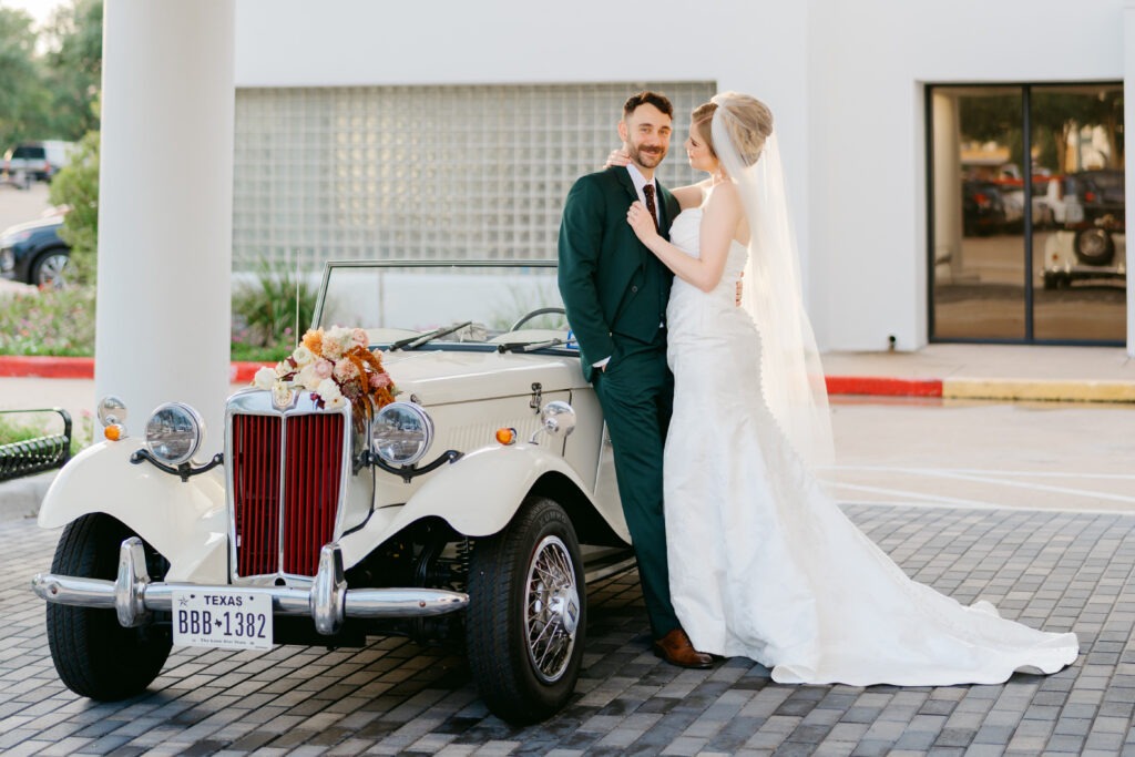 A newly married couple stare lovingly at each other as they lean against a vintage automobile. They are outside the Hilton hotel with the bright white building behind them. They are sheltered by the porte-coch&egrave;re. The setting gives the feeling of two people in love ready to drive away and start their lives together.