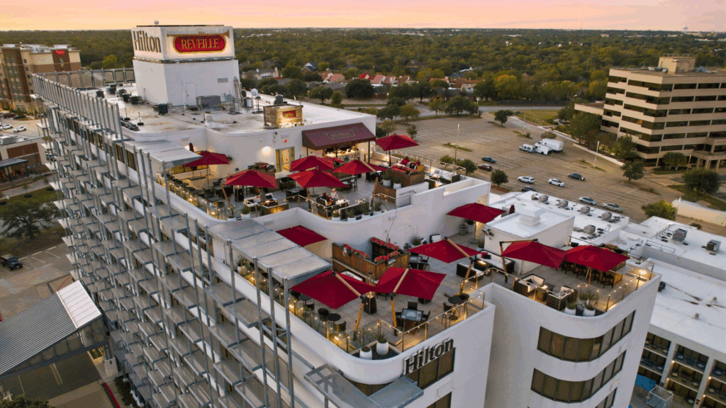 An aerial view of the Hilton Hotel with the rooftop bar, Reveille, featuring red umbrellas and seating areas. In the background, the city stretches out in the distance with roads, cars, and surrounding buildings visible. Above, the sky glows with vivid pink and orange streaks from the sunset, creating a striking backdrop over the urban landscape.